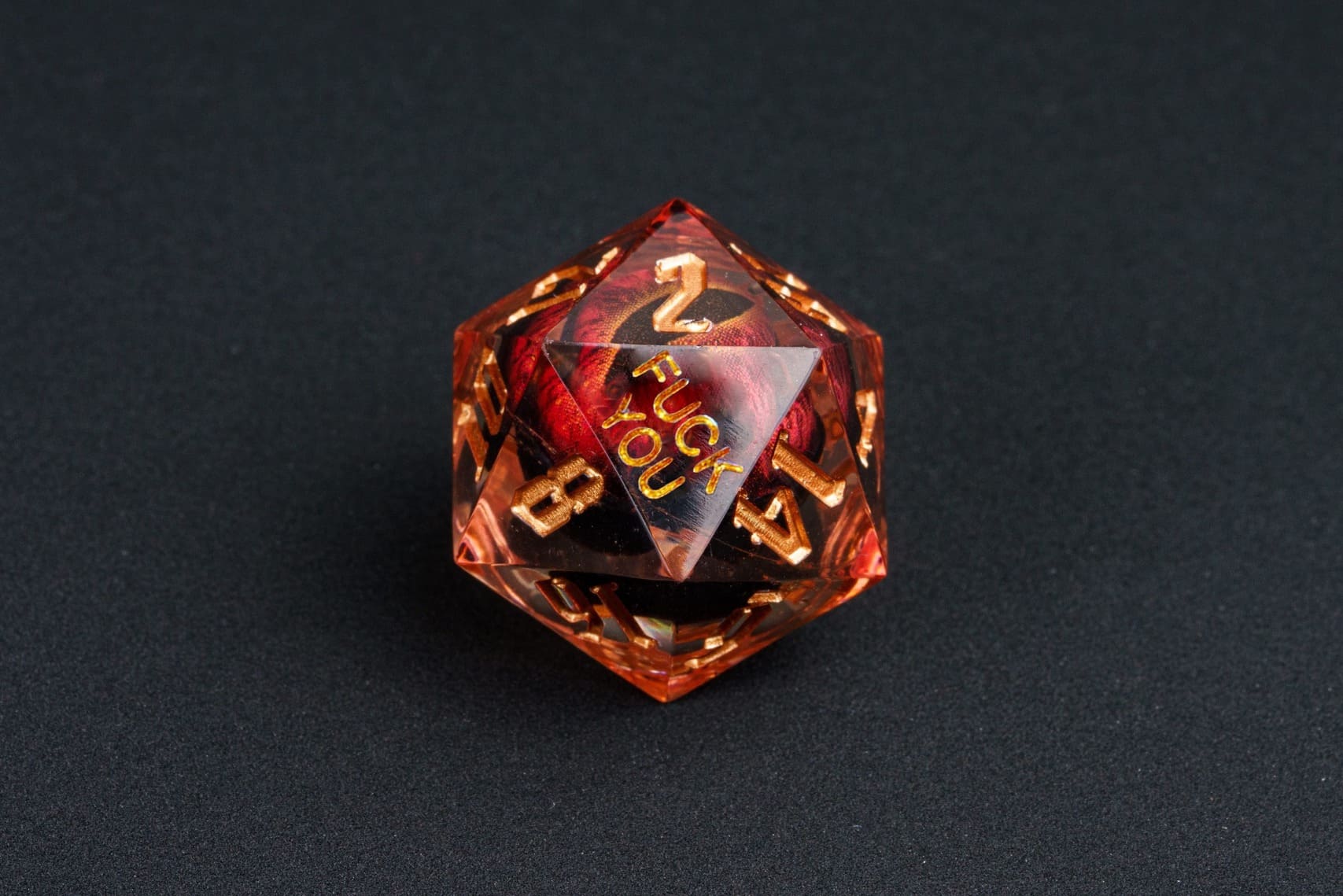 Close-up of the Dragon Eye Red D20 from the Fck Me Sharp Edge Dice Set, transparent red resin with fiery red swirl inside, gold numbering, and 'Fck You' lettering