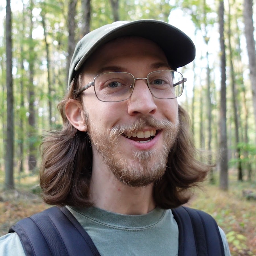 Smiling Bob World Builder with glasses and a green cap standing in a forest, wearing a backpack
