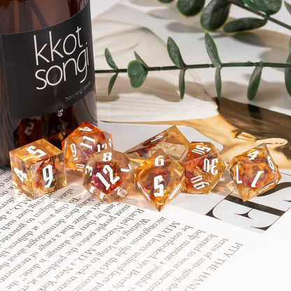 Aether Shards Sharp Edge Dice Set arranged on an open book beside a vase, highlighting translucent amber resin dice with gold foil inclusions