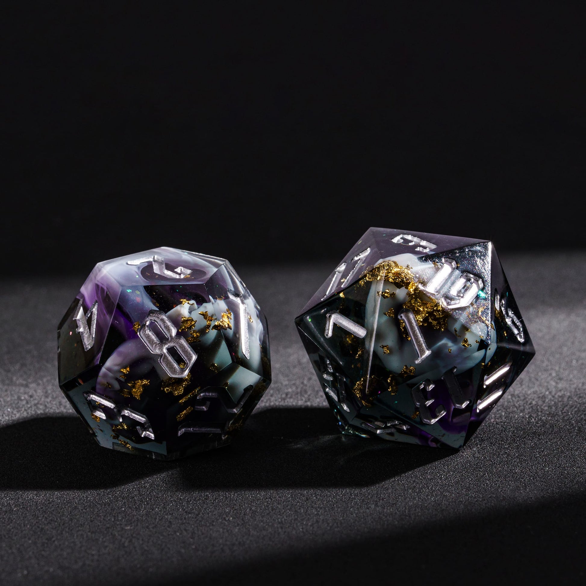 Close up of Siren’s Cache sharp edge D20 and D12 resin dice with gold foil flakes and silver numbering on black background