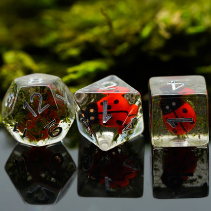 Trio of clear resin dice with red ladybugs inside, featuring D12, D8, and D6 on moss background